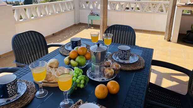 Una mesa lista para el desayuno, con frutas, pasteles y bebidas, con vistas a un paisaje marino.