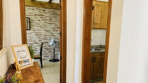 Una mesa de madera para comer con un menú enmarcado, un ventilador y una planta. Se ve una cocina con armarios de madera y un salpicadero de azulejos a través de un vano abierto.