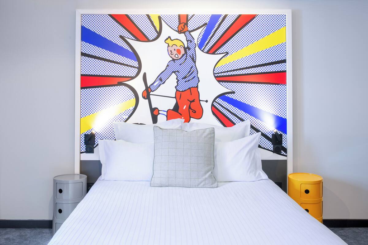 Een hotelkamer met een bed, kussens, een nachtkastje, een lamp en een kunstwerk aan de muur.