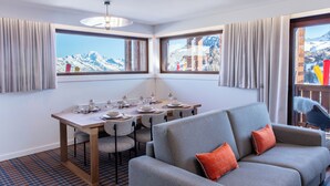 Une chambre d’hôtel moderne dotée d’un coin repas, d’un canapé gris et de grandes fenêtres offrant une vue sur les montagnes.