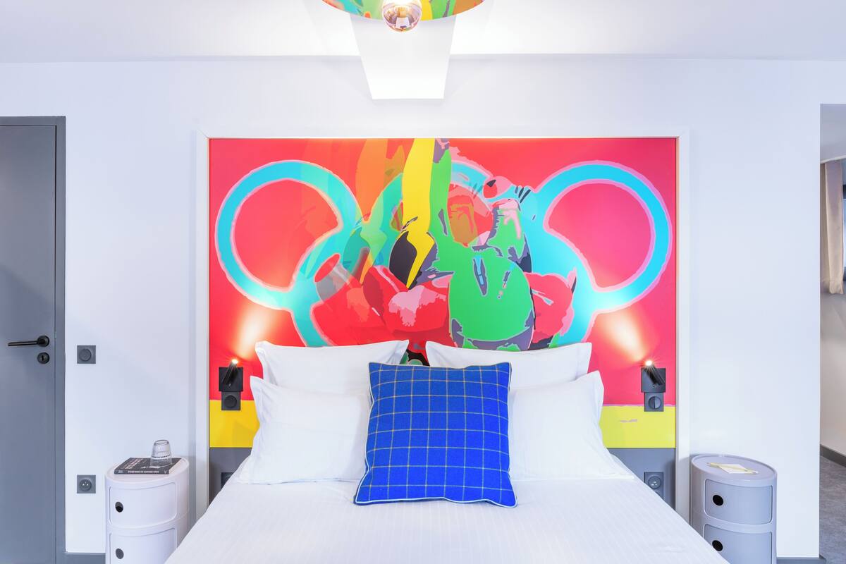 Een hotelkamer met een bed, kussens, een nachtkastje, een wandlamp en een kleurrijk kunstwerk aan de muur.