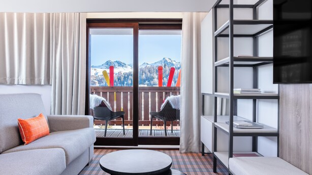 Une chambre d’hôtel moderne avec un canapé, une petite table ronde, une bibliothèque et un balcon offrant une vue sur les montagnes.