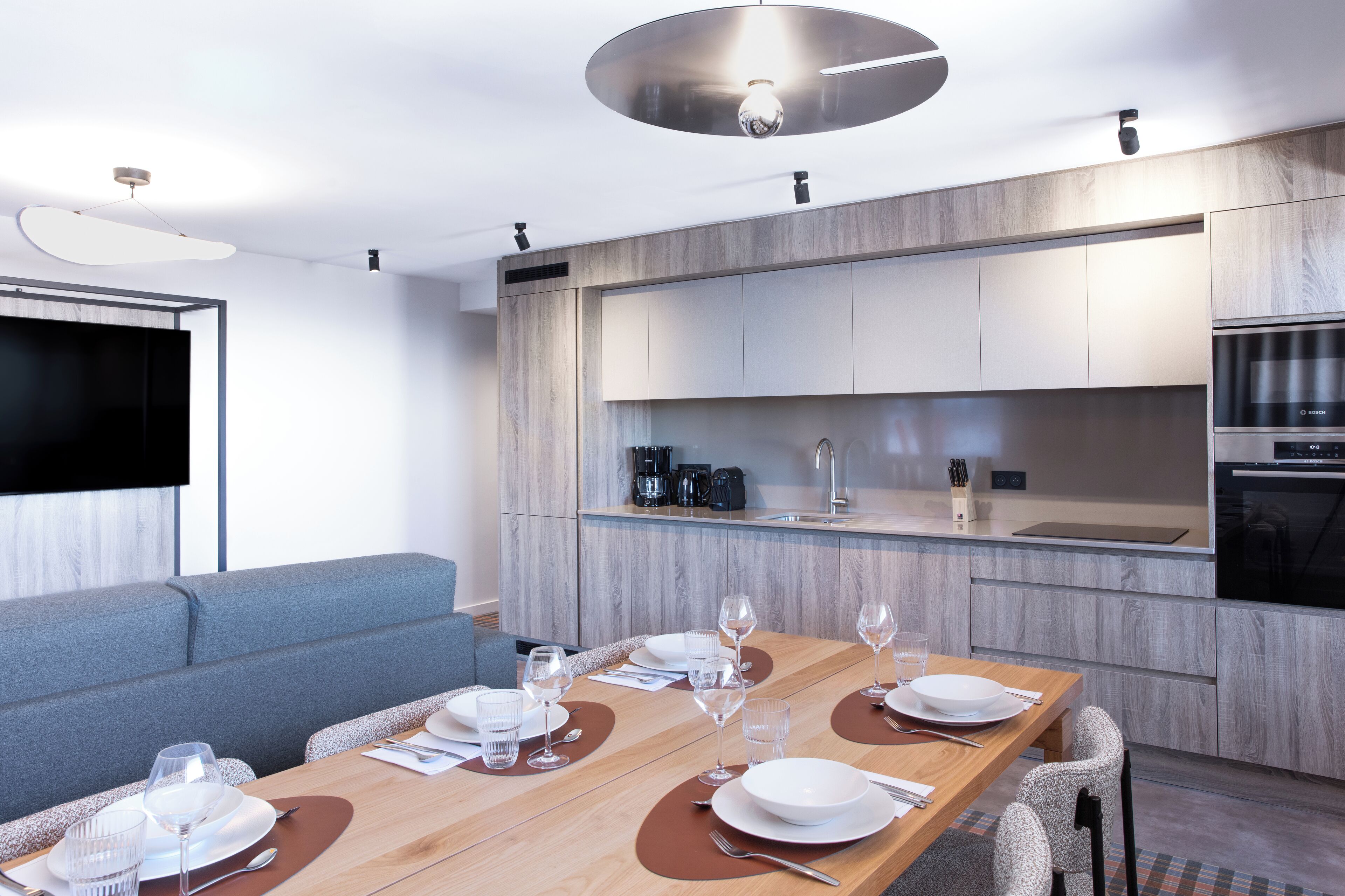 A modern kitchen with built-in appliances, a dining area with a wooden table set for six, and a grey sofa in the living area.
