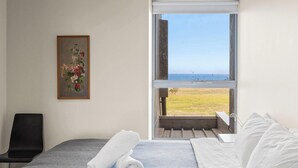 A bedroom with a bed, a rolled towel, a chair, a painting, and a view of the sea through an open window. - Ocean Views, 2 Bedroom Apartment (San Remo)