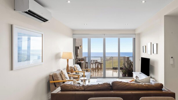 A modern living room with a sofa, chairs, a table, and a view of the sea through large glass doors. - Ocean Views, 2 Bedroom Apartment (San Remo)