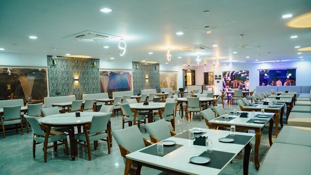 A spacious restaurant with multiple dining tables and chairs, a modern ceiling design, and a large mural on the back wall. - Sky View Resort (Bhuj)