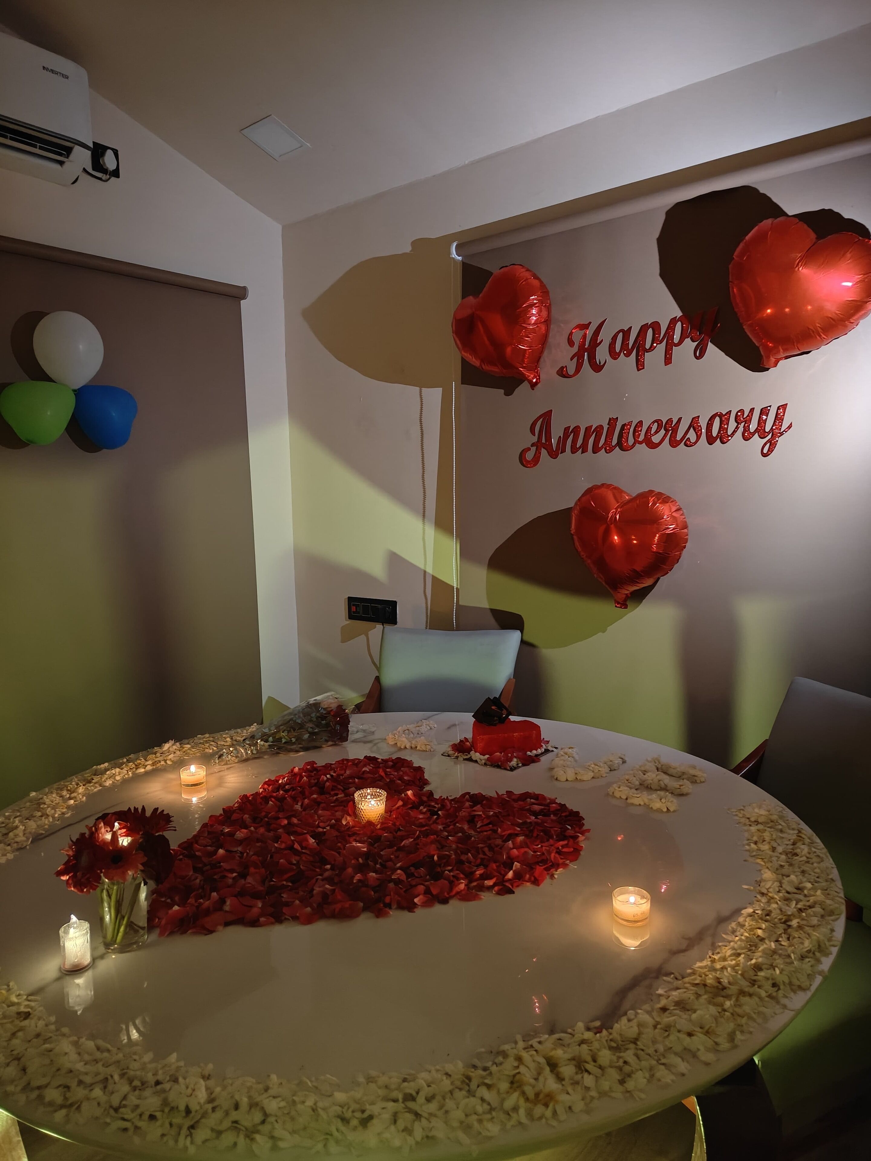 Una mesa redonda decorada con pétalos de rosa, velas y un pastel, rodeada de globos con forma de corazón y un cartel que dice 'Feliz aniversario'.