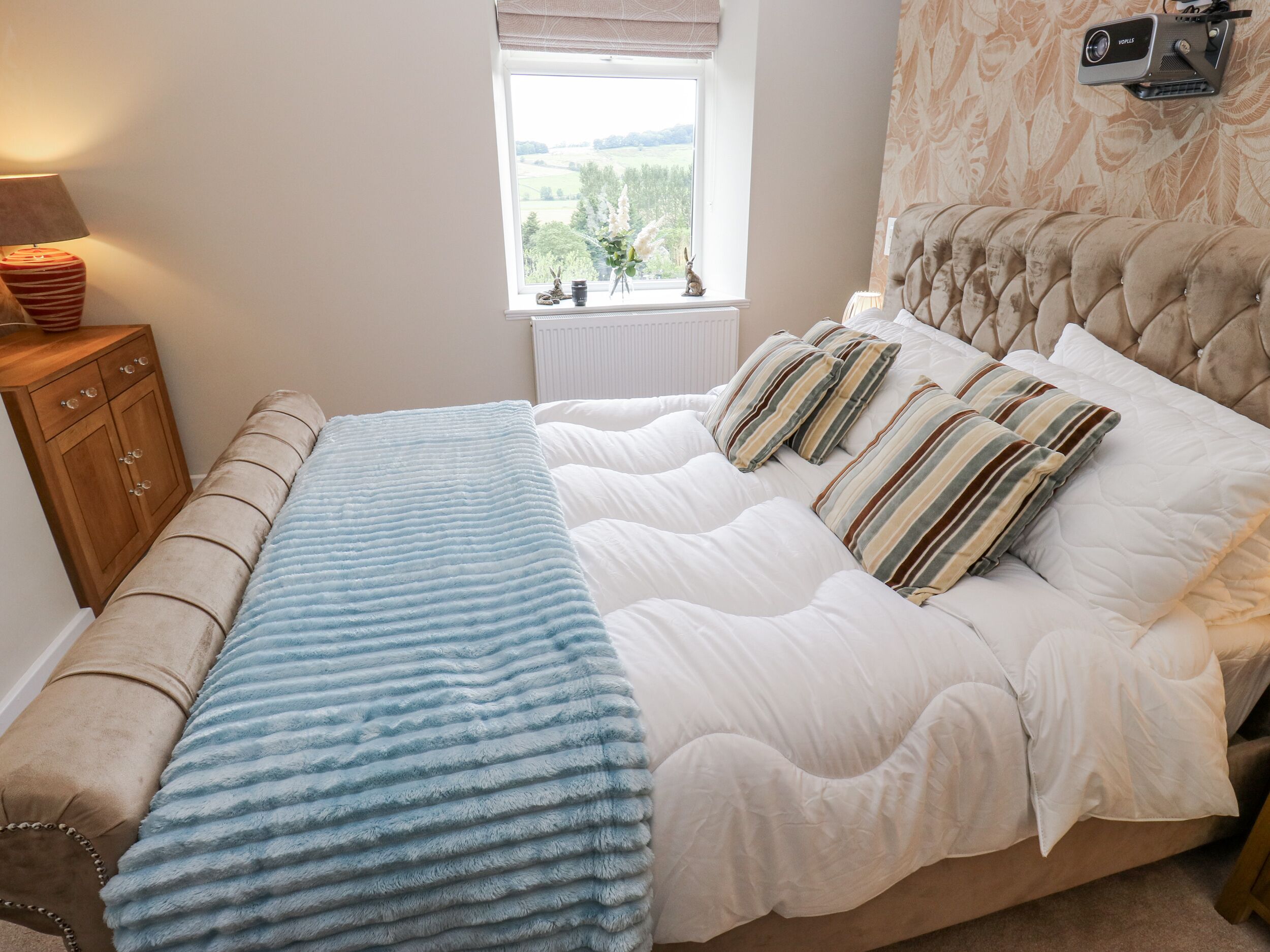 A bedroom with a bed, a bedside table, a lamp, a window, and a projector.