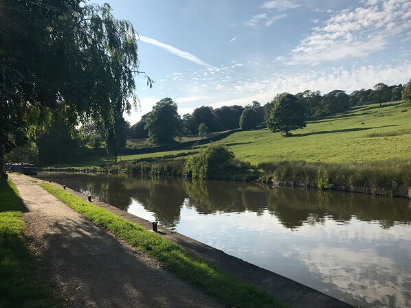 A tranquil riverside path with a calm water body, lush greenery, and trees in the background. - Millside View (Keighley)