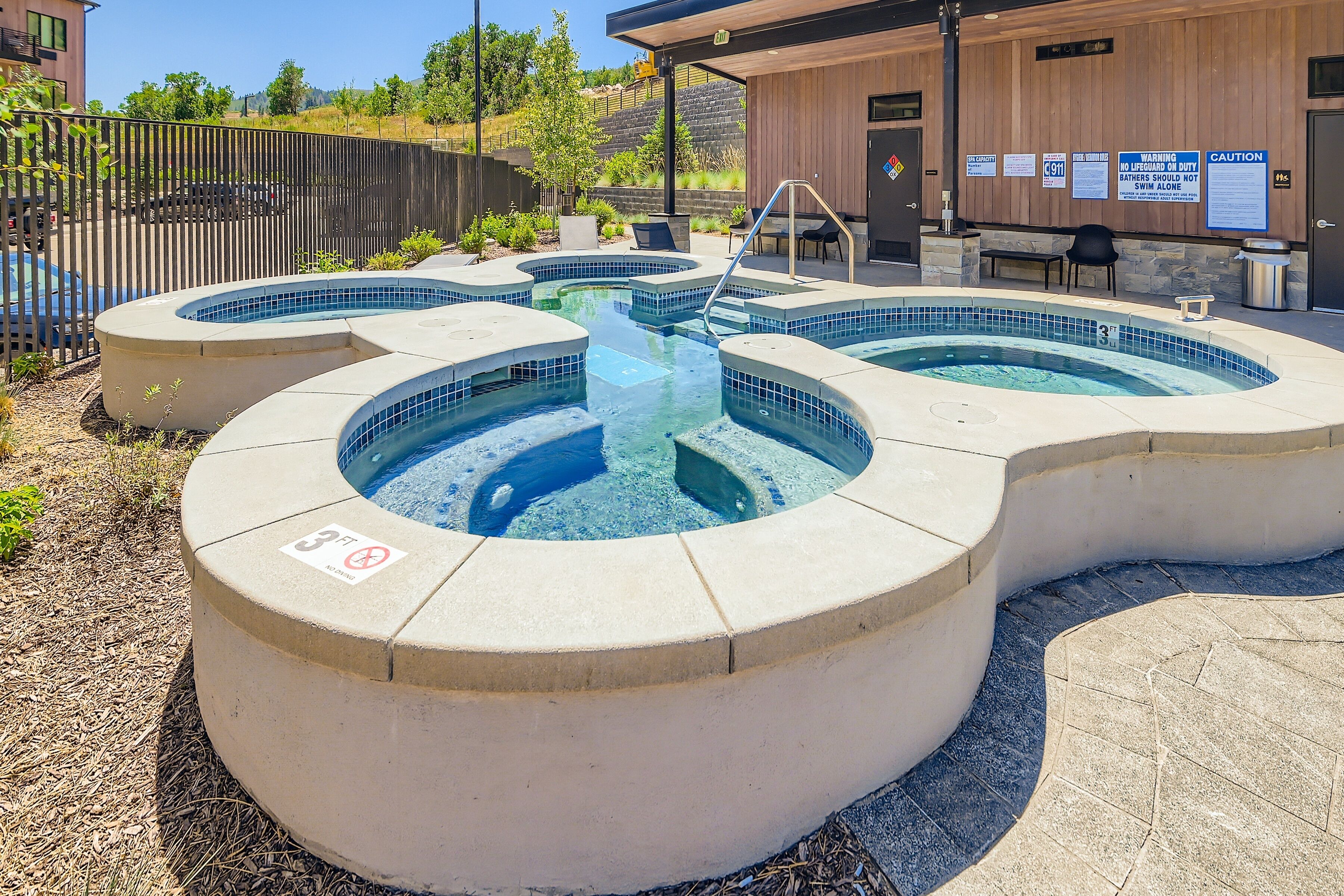 Un espace avec un jacuzzi, agrémenté d’une pancarte indiquant une profondeur minimale de 3 pieds pour éviter de plonger, d’une clôture en bois et d’un bâtiment avec une enseigne visible à l’arrière-plan.