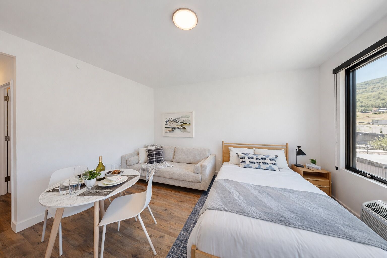 A modern, minimalist bedroom with a bed, a small table with chairs, a sofa, and a large window offering a view of the outdoors.