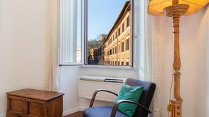 A room with a window, a radiator, a wooden table, a chair with a cushion, and a floor lamp. - Rental in Rome Casa Assunta (Rome)