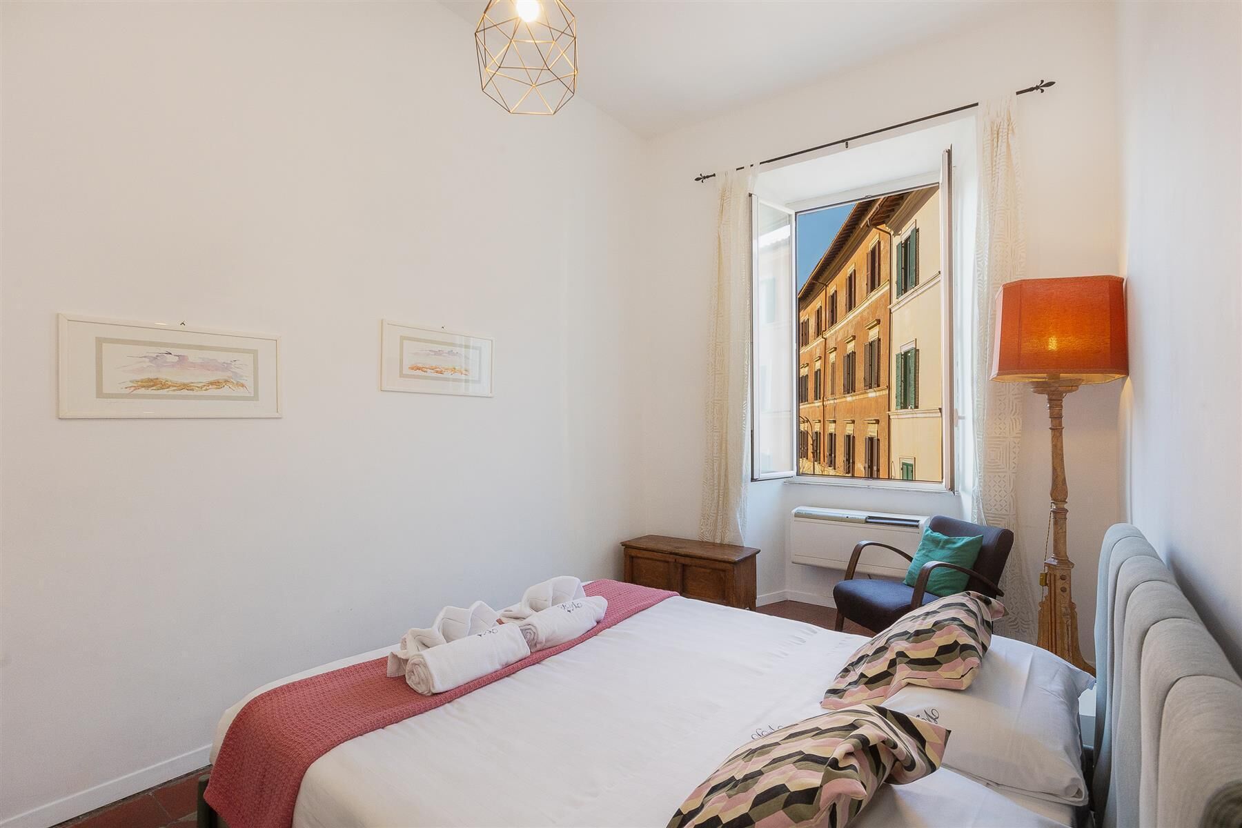 A hotel room with a bed, a window with a view of buildings, a bedside table, a lamp, and two framed pictures on the wall.