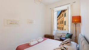 A hotel room with a bed, a window with a view of buildings, a bedside table, a lamp, and two framed pictures on the wall. - Rental in Rome Casa Assunta (Rome)