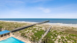 Un hotel frente a la playa con un muelle largo que se adentra en el océano, piscina y zona de playa con tumbonas.