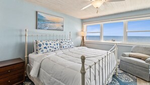 Un dormitorio con cama, mesita de noche, silla, ventilador de techo y ventana con vista al océano.