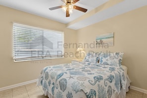 A bedroom with a bed, a ceiling fan, a window with blinds, a nightstand with a lamp, and a painting on the wall. - 2622 Forest Ridge #c2 3 Bedroom Condo (Fernandina Beach)