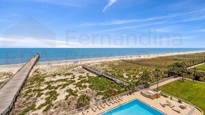 A beach view with a wooden pier, a swimming pool, and a fenced area with lounge chairs. - 667 Amelia By The Sea 3 Bedroom Condo (Fernandina Beach)