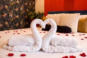 A bed with a heart-shaped towel arrangement, a floral-patterned wall, and a red and white pillow.