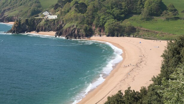 Paisaje costero con playa de arena, costa rocosa y una colina con vegetación.