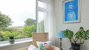 Una silla de mimbre con un cojín colorido y un libro, una lámpara azul y una planta en maceta con hojas grandes junto a una ventana con vistas a la vegetación.