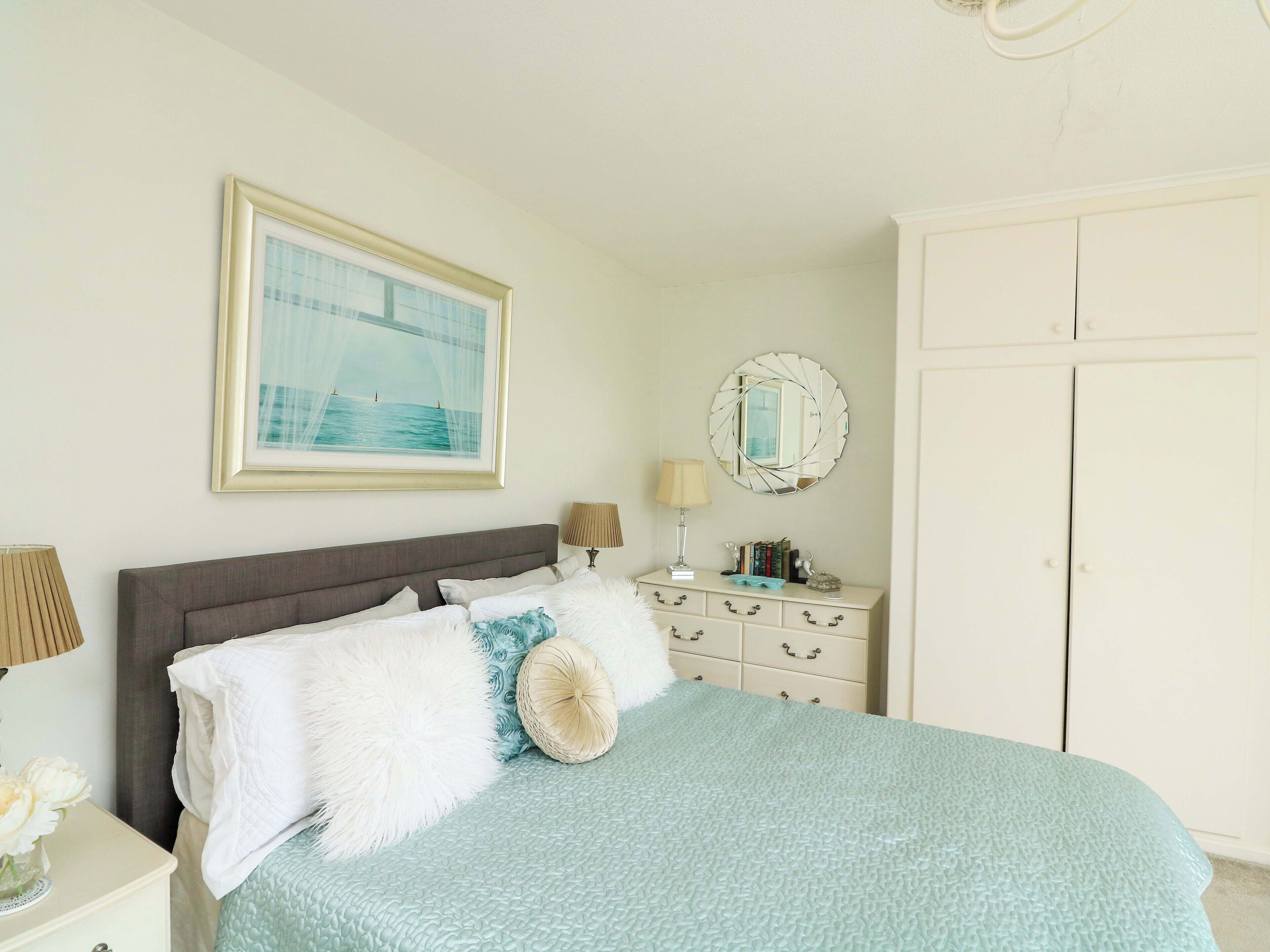 Une chambre à coucher bien rangée, avec un lit, une table de chevet dotée d’une lampe, une commode, un miroir et une armoire.
