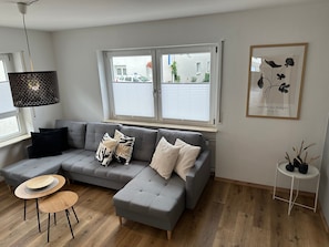 A modern living room with a grey sectional sofa, a round wooden table, and a small white side table with decorative items. - Apartment 'Groß' with Shared Garden, Balcony and Wi-Fi (Appenweier)