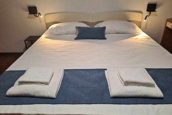 Una cama bien hecha con sábanas blancas, un plaid azul y dos toallas blancas dobladas.