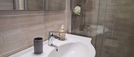Un baño con un lavamanos blanco, un grifo cromado, dispensador de jabón, una lata de refresco Cherry y una zona de ducha con puerta de vidrio.