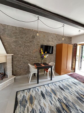 A modern living room with a stone wall, a TV mounted above a wooden cabinet, and a dining area with white chairs and a black table.