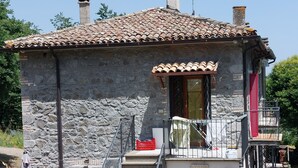 A stone building with a tiled roof, a staircase leading to a balcony, and a small garden area with potted plants.