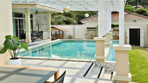 Un área de piscina con techo de cristal, una sala de estar con sillones y un edificio pequeño al fondo.