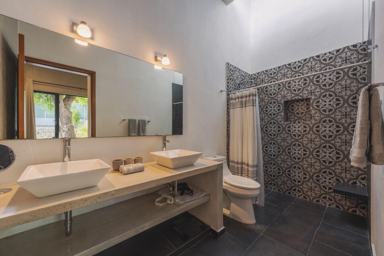 A modern bathroom with a large mirror, two sinks, a toilet, and a shower area with patterned tiles.