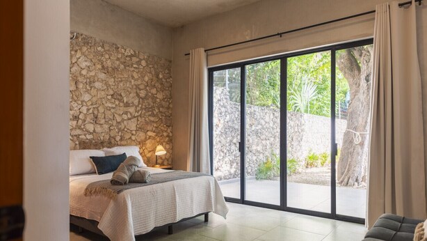 A bedroom with a stone wall, a bed with a blanket, a bedside lamp, and a view of a garden through a sliding glass door. - Casa Péepen Merida Centro with Pool by LAHOS (Mérida)