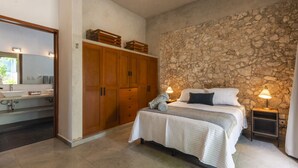 A bedroom with a stone wall, a bed with white bedding, a nightstand with a lamp, and a wardrobe with wooden doors. - Casa Péepen Merida Centro with Pool by LAHOS (Mérida)