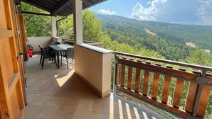 Un espacio exterior techado con una mesa, sillas, barandal de madera y vistas a un paisaje boscoso.