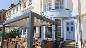 A building with a white facade, a blue door, and a covered outdoor seating area with tables and chairs. - The Beach Condo (Weymouth)
