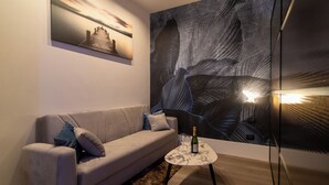 Una sala de estar moderna con un sofá gris, una mesita redonda con una botella y un vaso, y un cuadro abstracto grande en la pared.