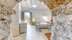 Un dormitorio con pared de piedra, cama con cabecera verde, mesita de noche, una silla y una ventana con cortinas.