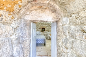 An interior view of a stone-walled room with a bed, a table, and a chair.