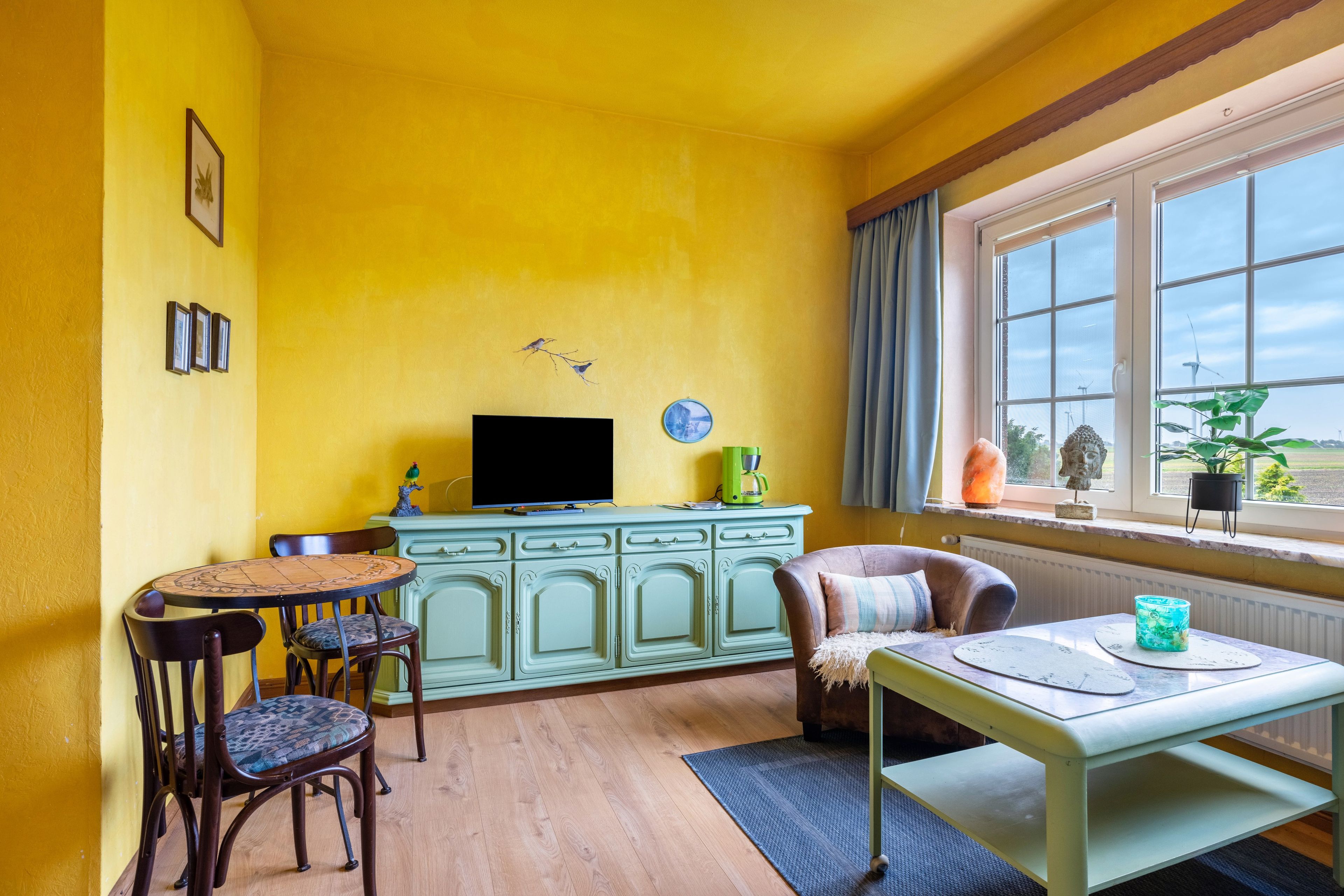 Ein Zimmer mit gelben Wänden, Holzboden, einem Esstisch mit Stühlen, einem Fernseher auf einer Kommode und einem Fenster mit Blick auf Windräder.