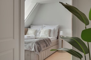 A bedroom with a sloped ceiling, a bed with white bedding, a bedside table with a lamp, and a potted plant.