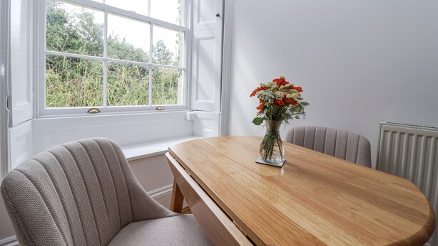 Un comedor con mesa de madera, dos sillas tapizadas, un florero con flores y una ventana con vistas a la vegetación.