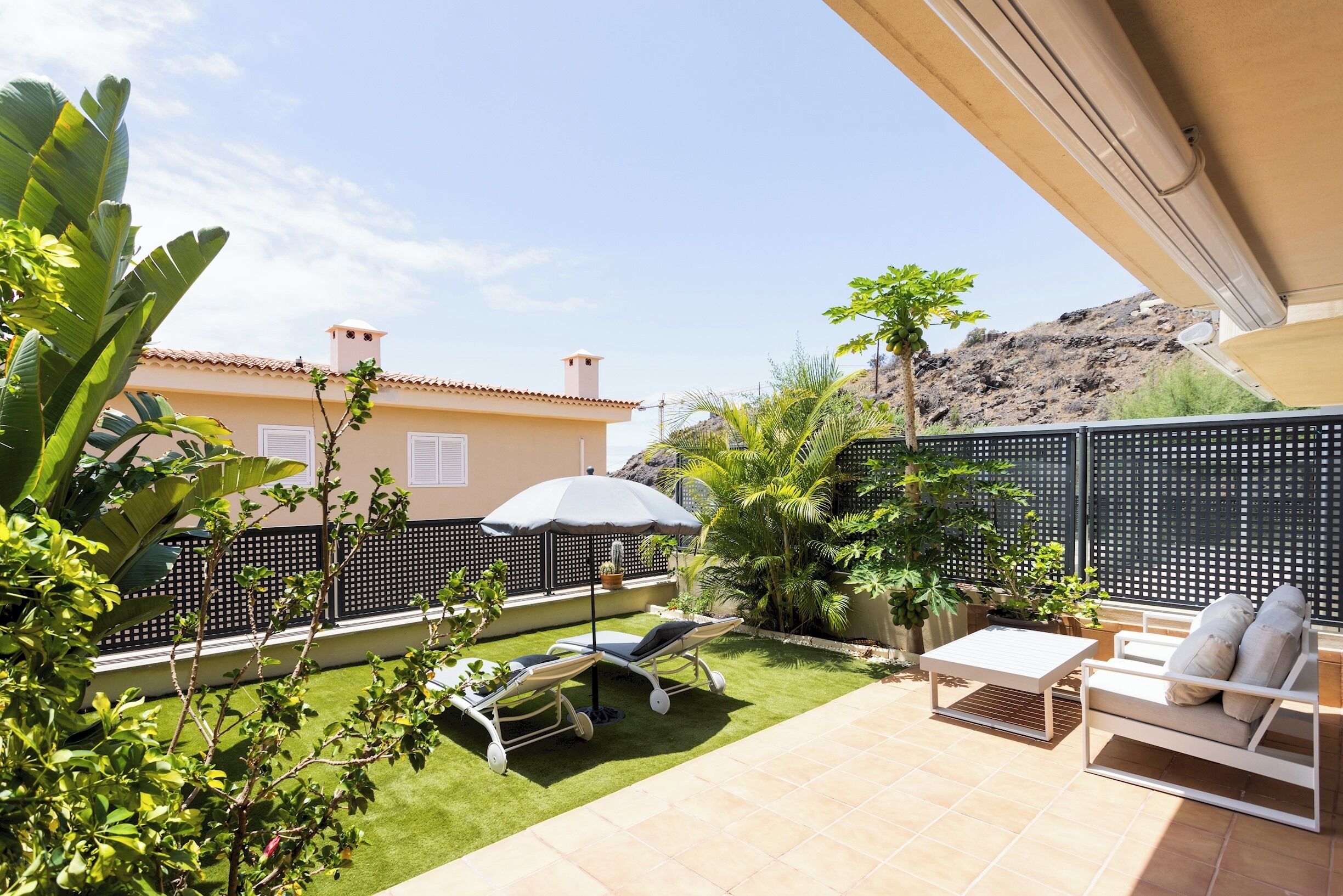 Un patio con dos sillones de descanso, una mesita y una sombrilla. Se observa una cerca y abundante vegetación.
