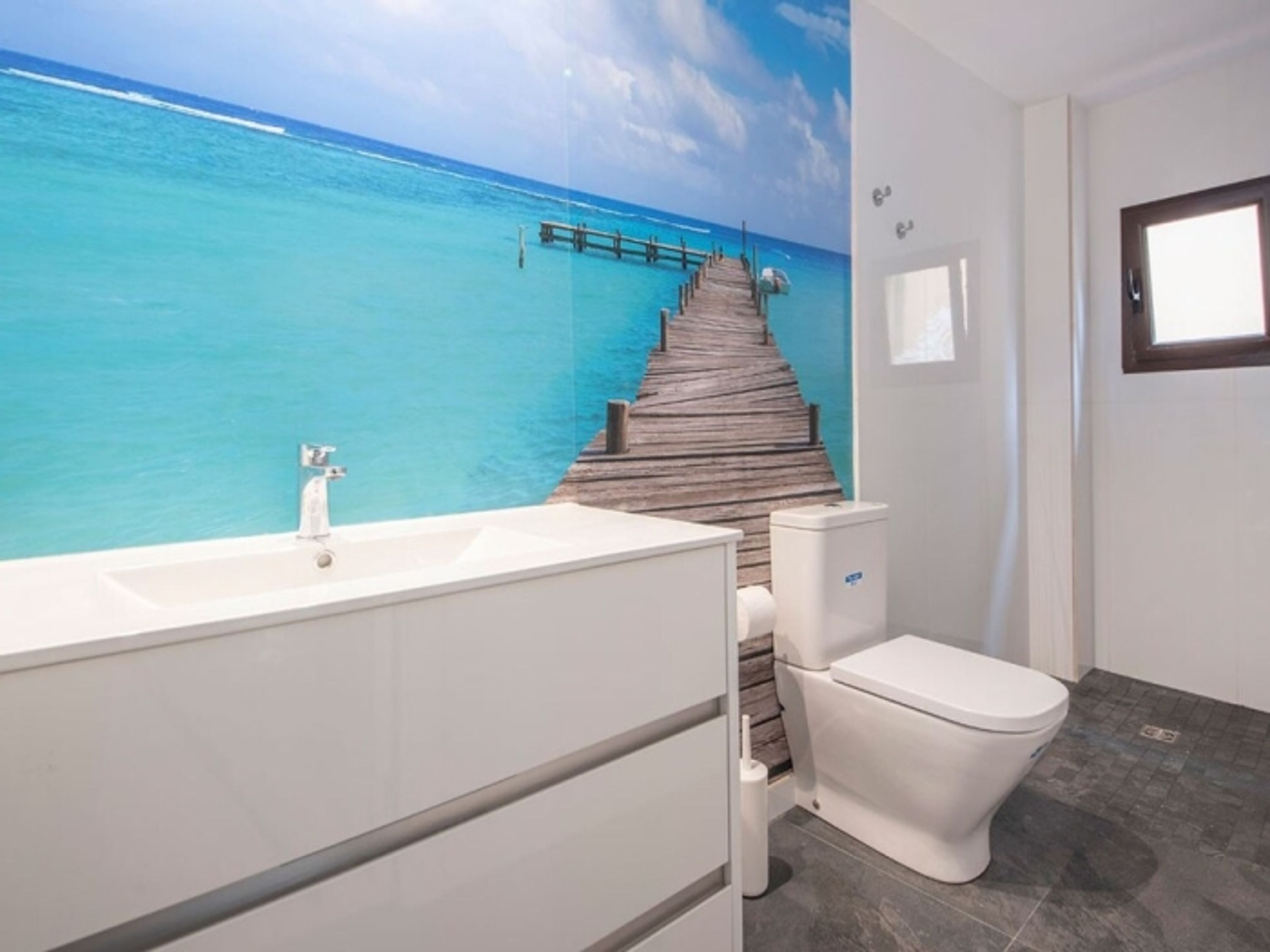 A bathroom with a wooden dock leading to a turquoise sea, a white sink, and a toilet.