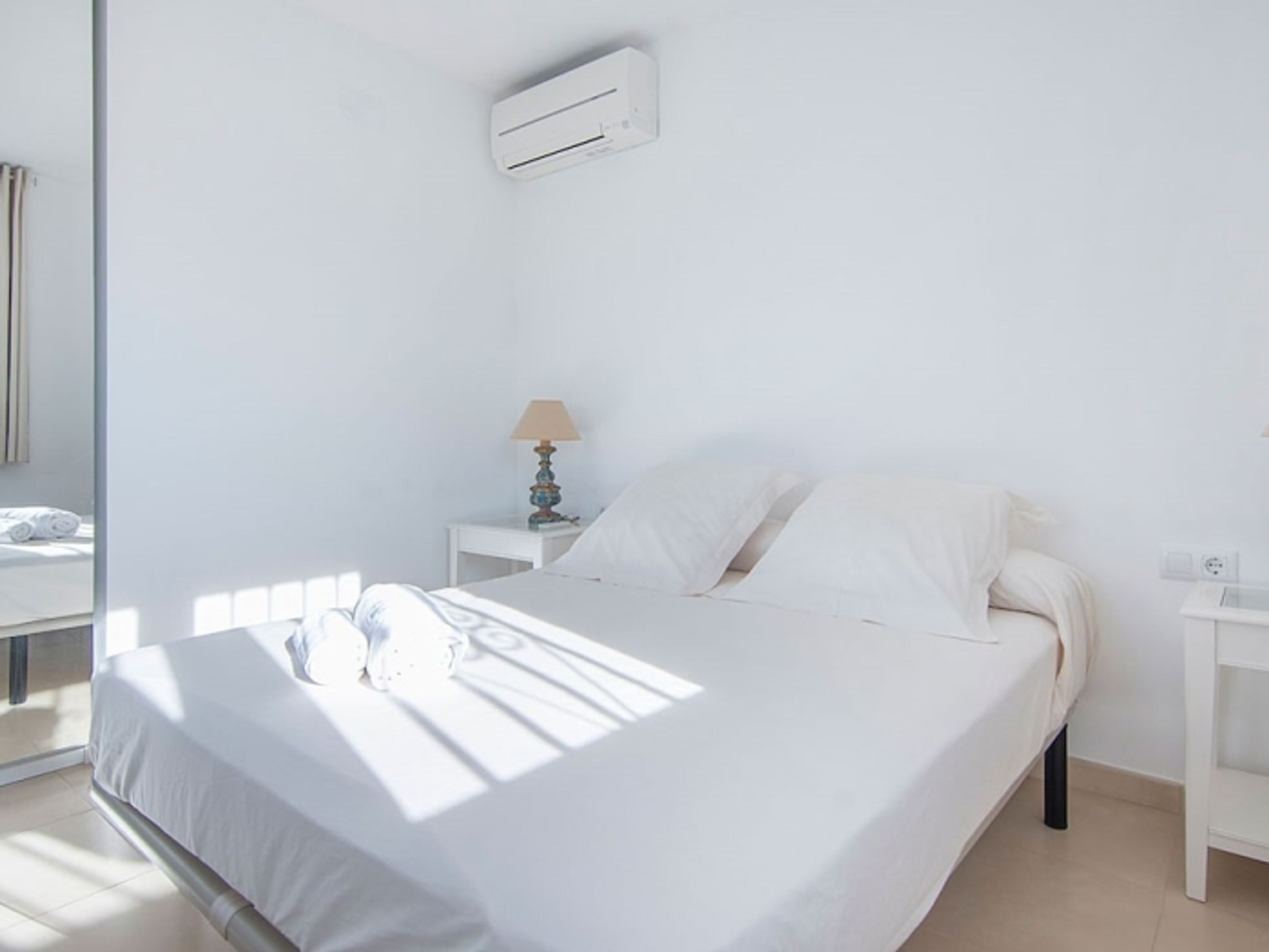 Una cama bien hecha con sábanas blancas, una lámpara de noche y un equipo de aire acondicionado en la pared.