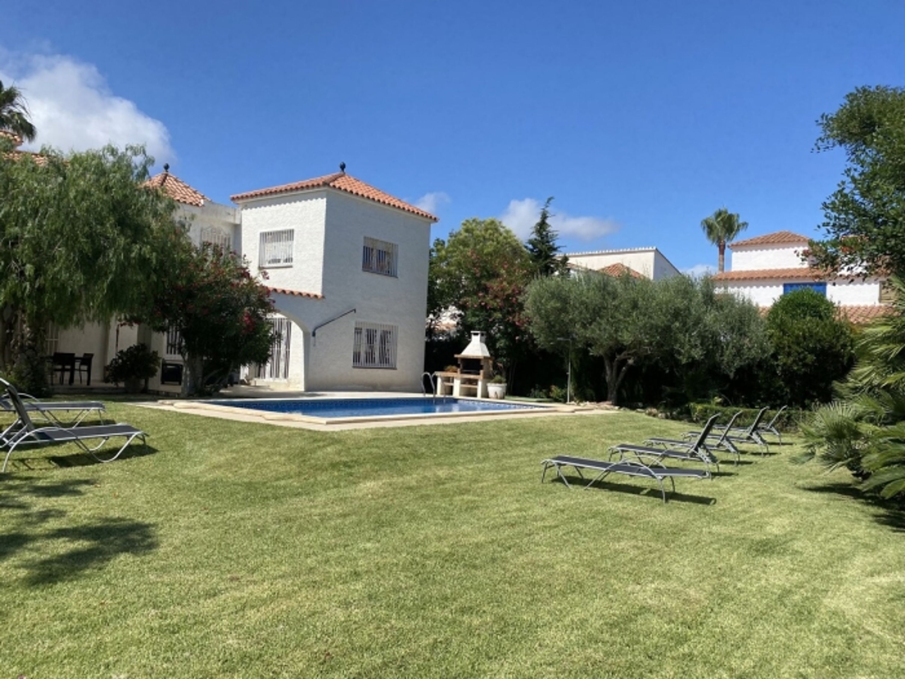 Un área de piscina con sillones de descanso, un pequeño edificio con tejado de tejas rojas y vegetación circundante.