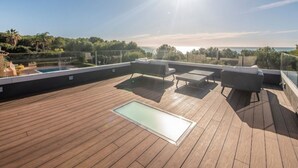 Een dak terras met een houten dek, buiten zitplaatsen en een glazen hekwerk met uitzicht op een kustlandschap.