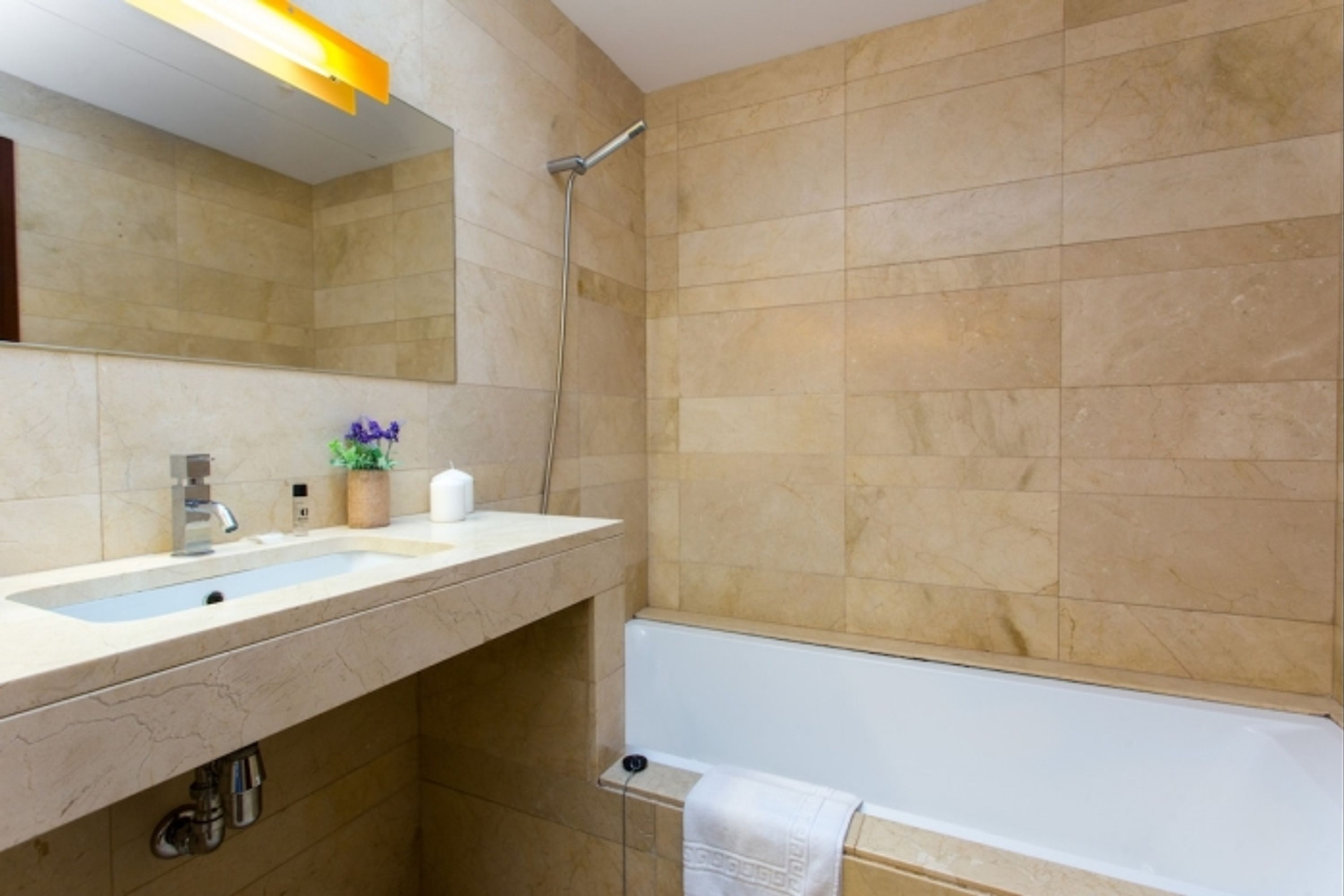 Ein modernes Badezimmer mit einer beige gefliesten Wand, einem weißen Waschbecken mit Spiegel darüber, einer Handbrause und einer weißen Badewanne.