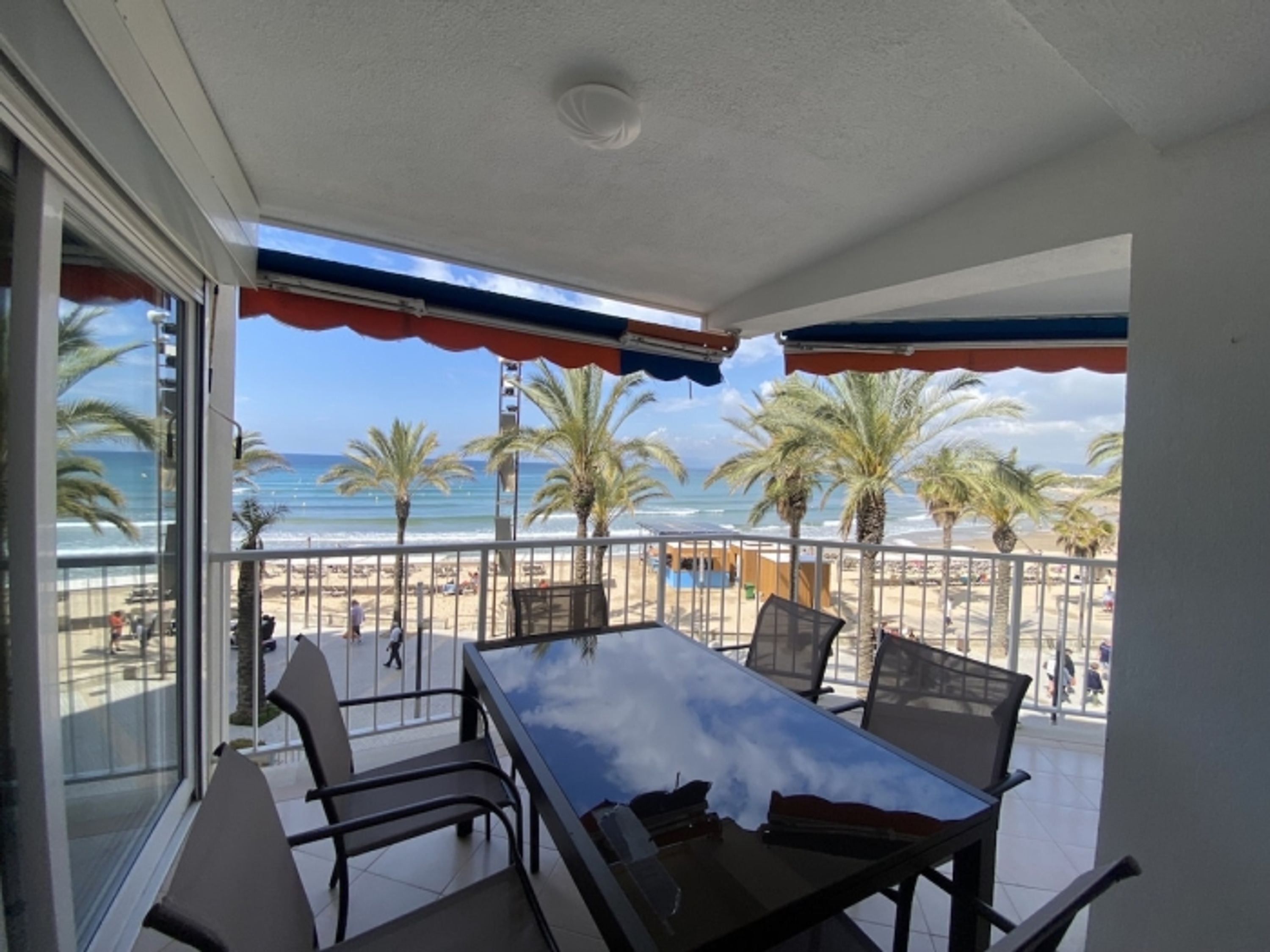 A balcony with a glass table and chairs, overlooking a beach with palm trees and the ocean.